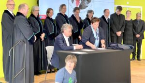 Dr. Richard Griffioen sits at a table and signs a paper. In the background we see lined up academia, dressed in black robes.