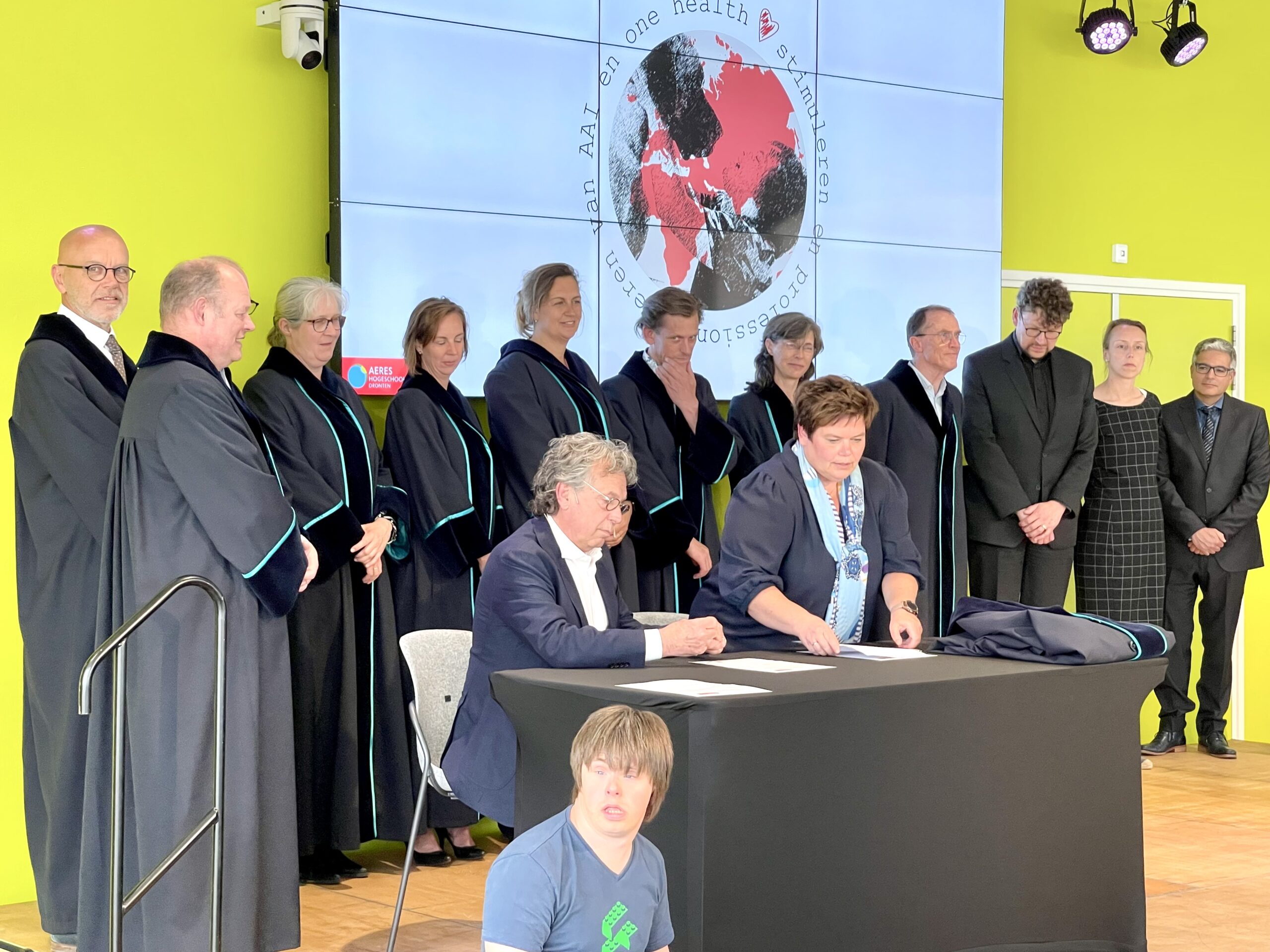 Dr. Richard Griffioen sits at a table and signs a paper. In the background we see lined up academia, dressed in black robes.